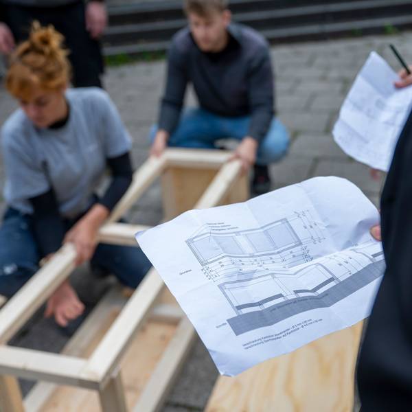 Im Vordergrund ist ein Bauplan der Sitzelemente für den Ebertplatz. Im Hintergrund sieht man verschwommen, wie sie gebaut werden.