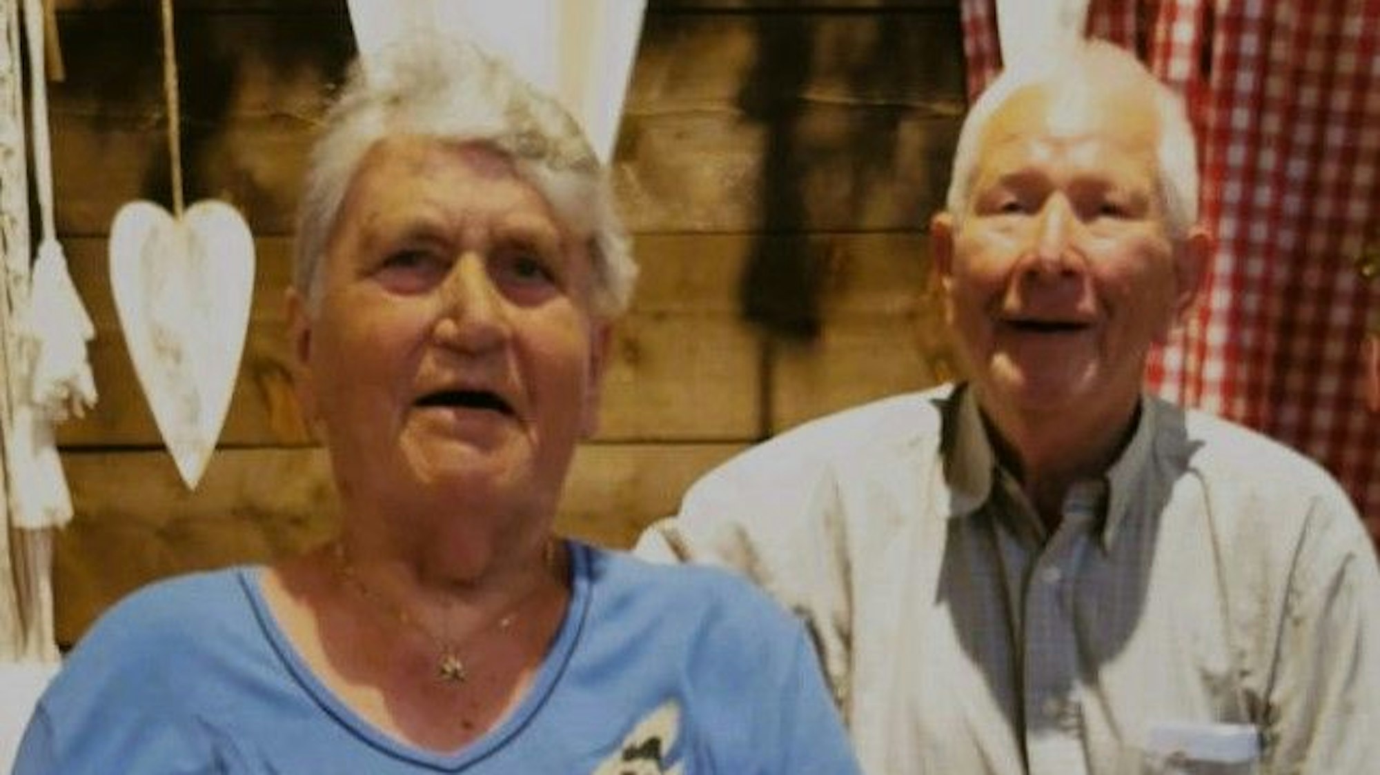 Das Paar schaut in die Kamera, die Wand hinter ihnen ist mit Herzen dekoriert.