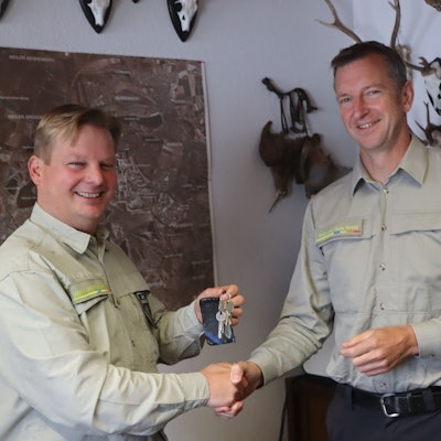 Zwei Männer schütteln sich vor einer Wand mit Landkarte und Jagdtrophäen die Hand.