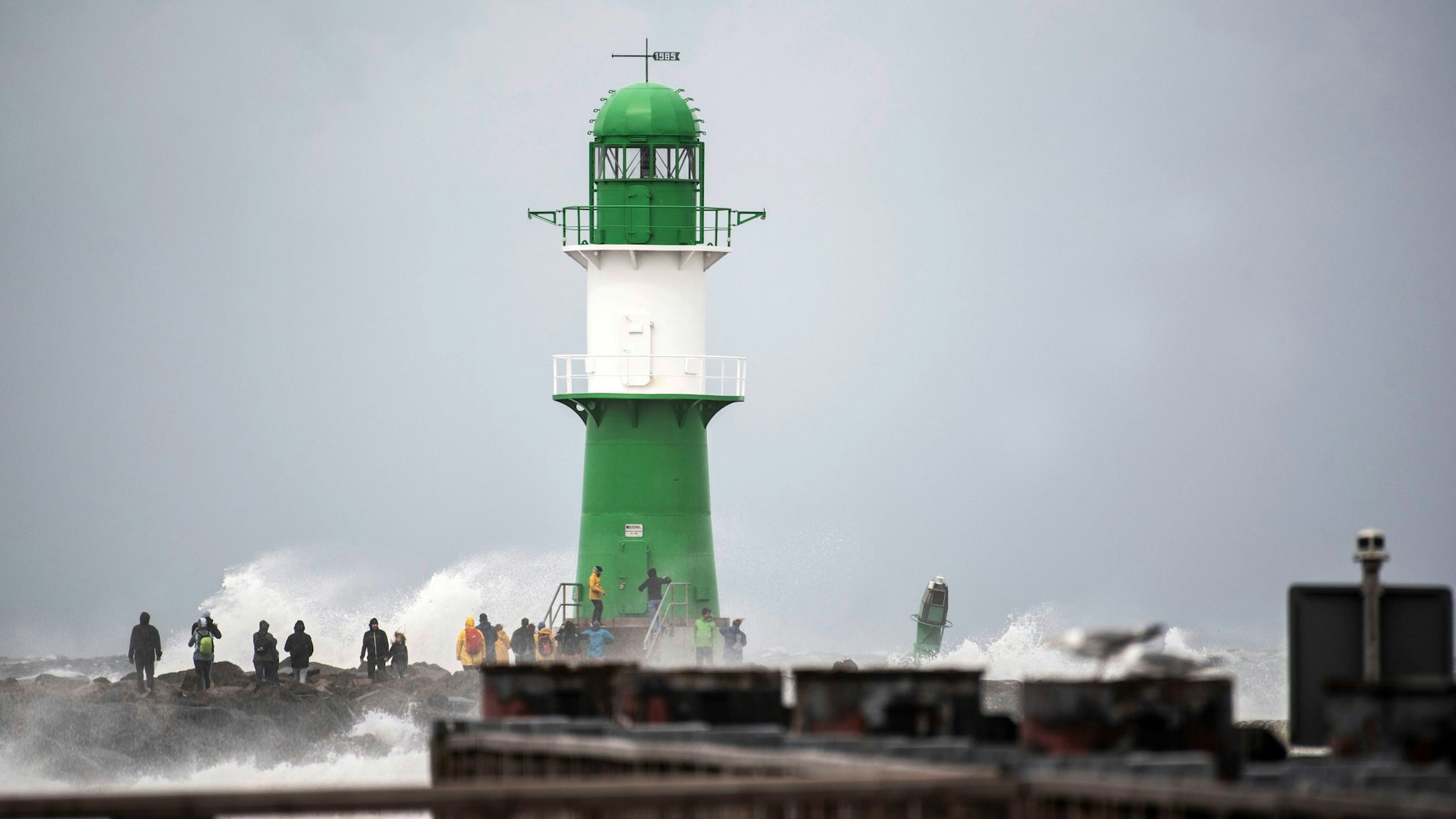 Schwerer Wellengang durch das Sturmtief „Zacharias“ an der Ostseeküste. Mehrere Schaulustige beobachten, wie die Wellen neben einem Leuchtturm bei Warnemünde an die Küste klatschen.