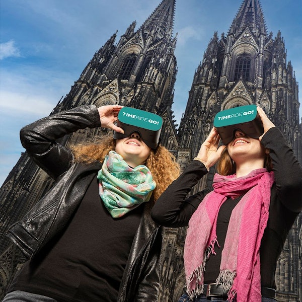 Zwei Frauen stehen vor dem Kölner Dom und schauen durch VR-Brillen mit der Aufschrift „Time Ride Go“