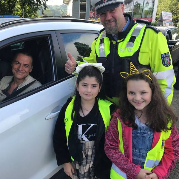 Ein Polizeibeamter in Uniform, zwei Grundschülerinnen und ein Autofahrer