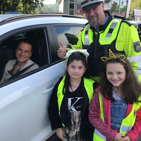 Ein Polizeibeamter in Uniform, zwei Grundschülerinnen und ein Autofahrer