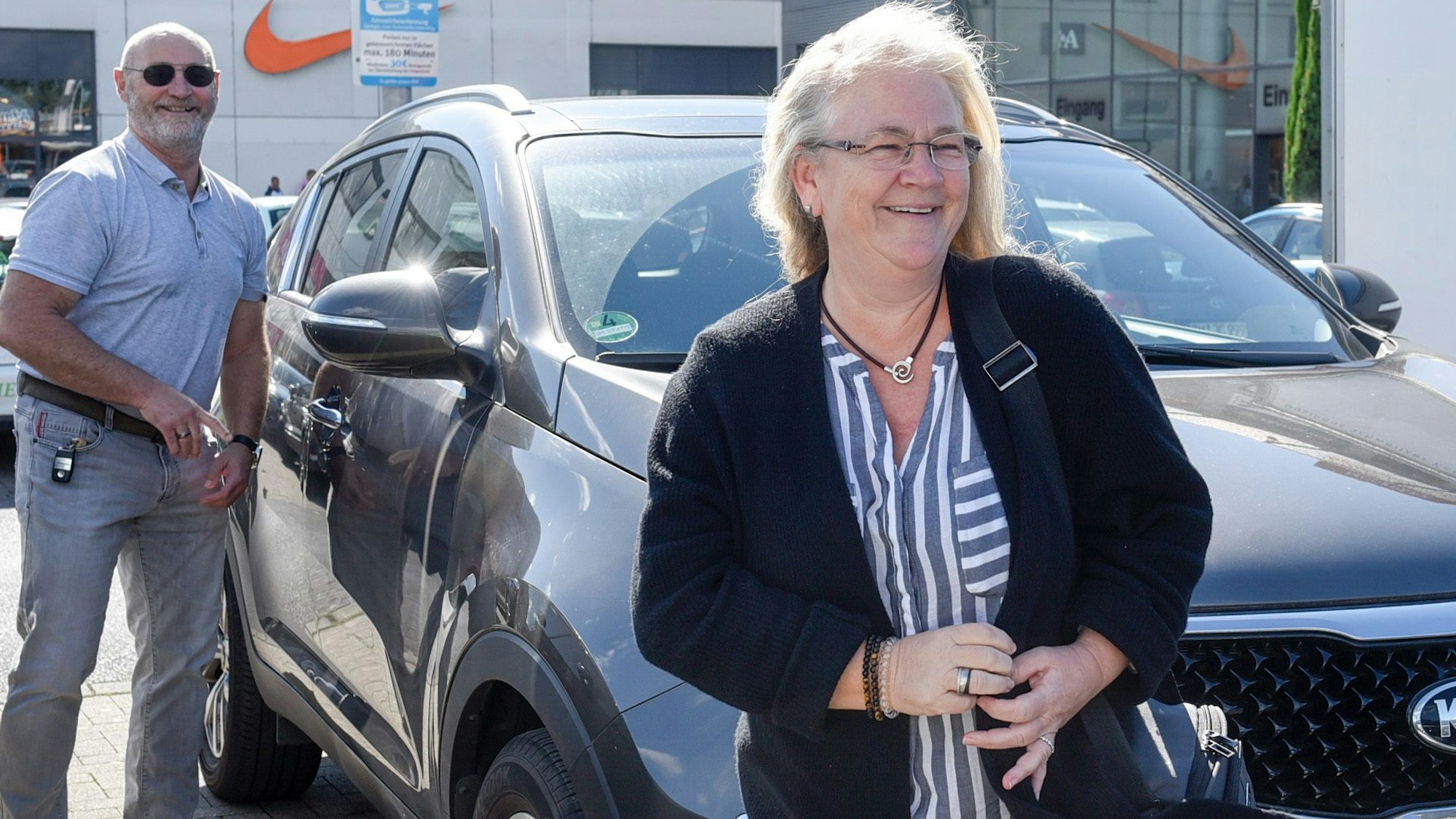 Das Bild zeigt das Ehepaar Lehnen an ihrem Auto auf dem Parkplatz des Einkaufszentrums.