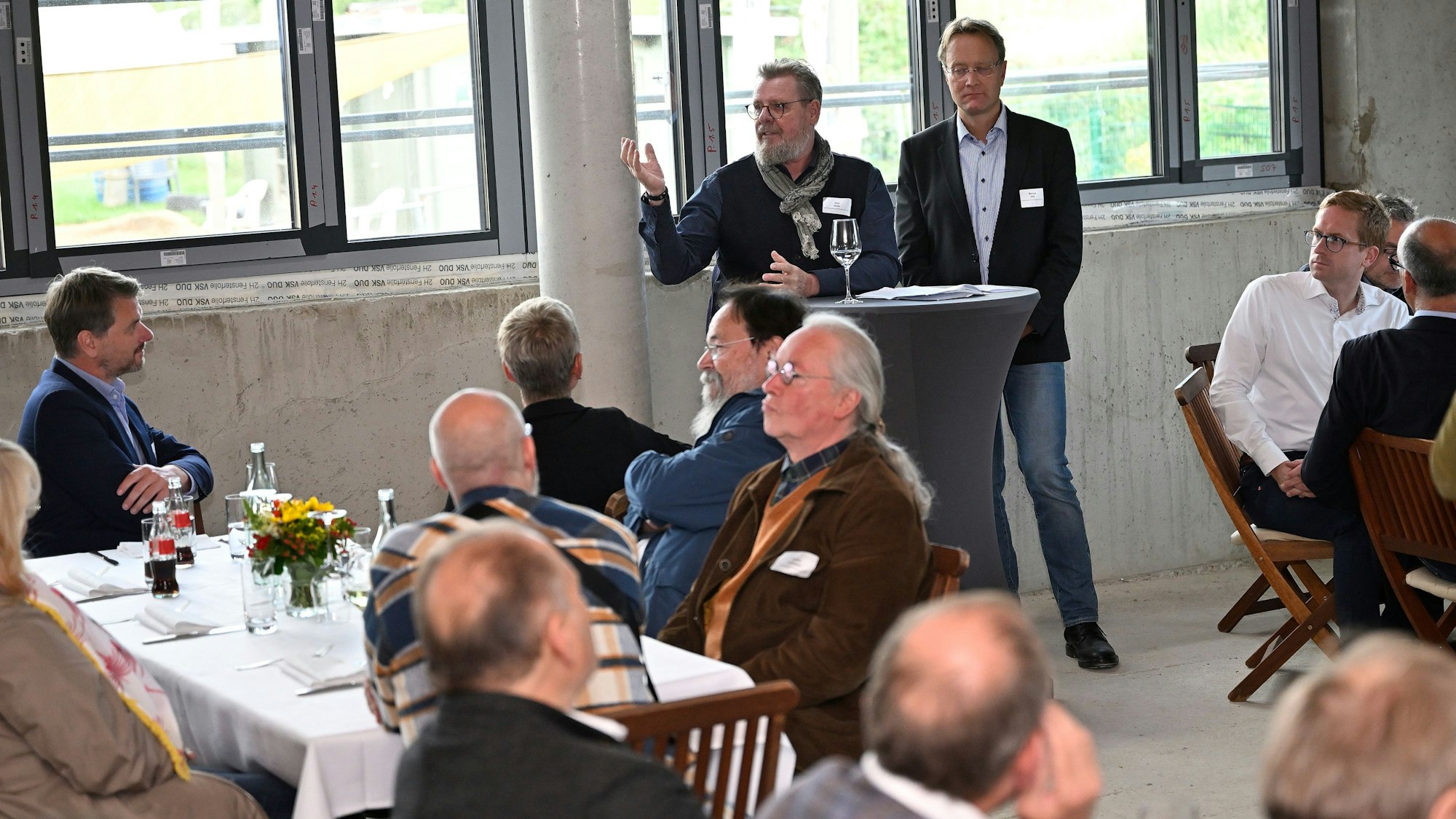 Zwei Männer stehen an einem Stehtisch, Gäste sitzen an Tischen mit weißen Tischdecken in einem Rohbau.