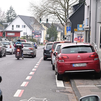 Zu sehen sind zwei Fahrspuren mit einem Rückstau vor der Ampelkreuzung. Der Radstreifen wird durch ein parkendes Auto blockiert. Der Parkstreifen ist vollständig belegt mit parkenden Autos.