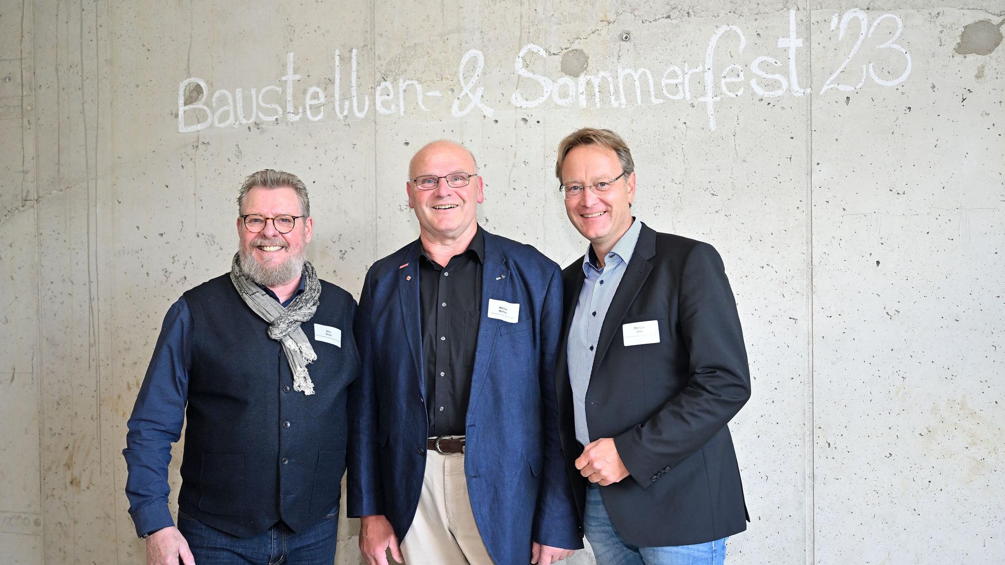 Drei Männer stehen vor einer Wand auf der Baustellen- und Sommerfest '23 steht.
