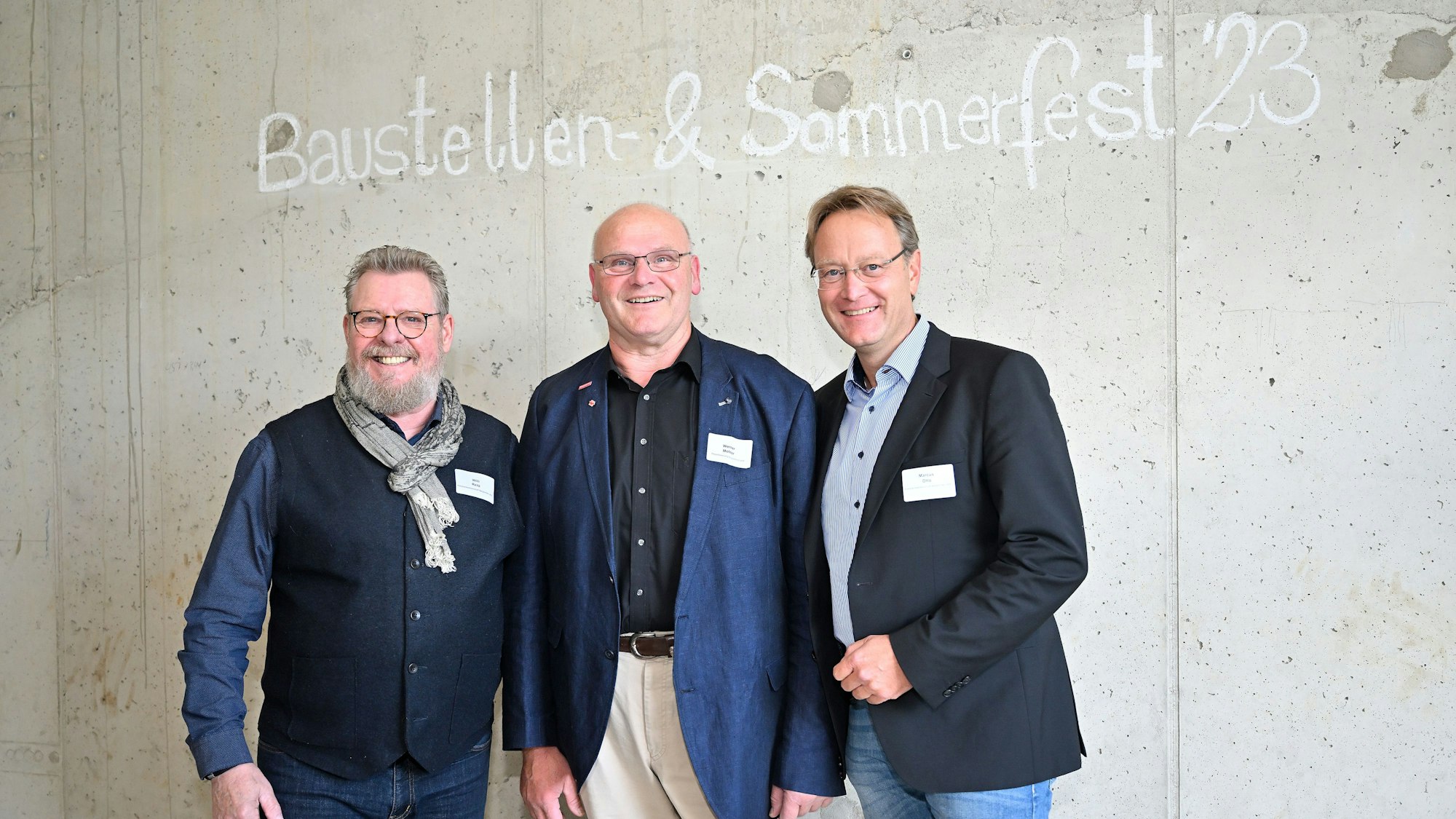 Drei Männer stehen vor einer Wand auf der Baustellen- und Sommerfest '23 steht.