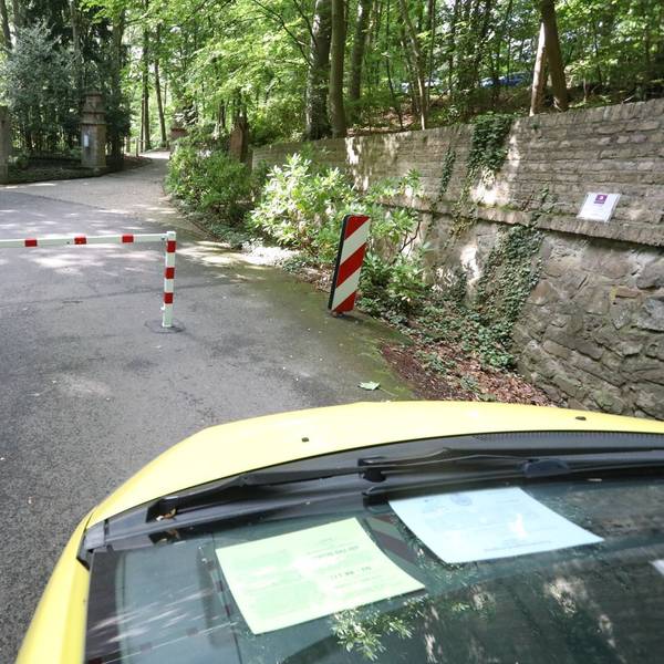 Die Front eines Autos ist im Bild, das vor einer rot-weiß gestreiften Sperre steht. Dahinter ist eine Straße in Wald zu erkennen.