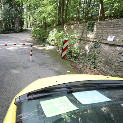 Die Front eines Autos ist im Bild, das vor einer rot-weiß gestreiften Sperre steht. Dahinter ist eine Straße in Wald zu erkennen.
