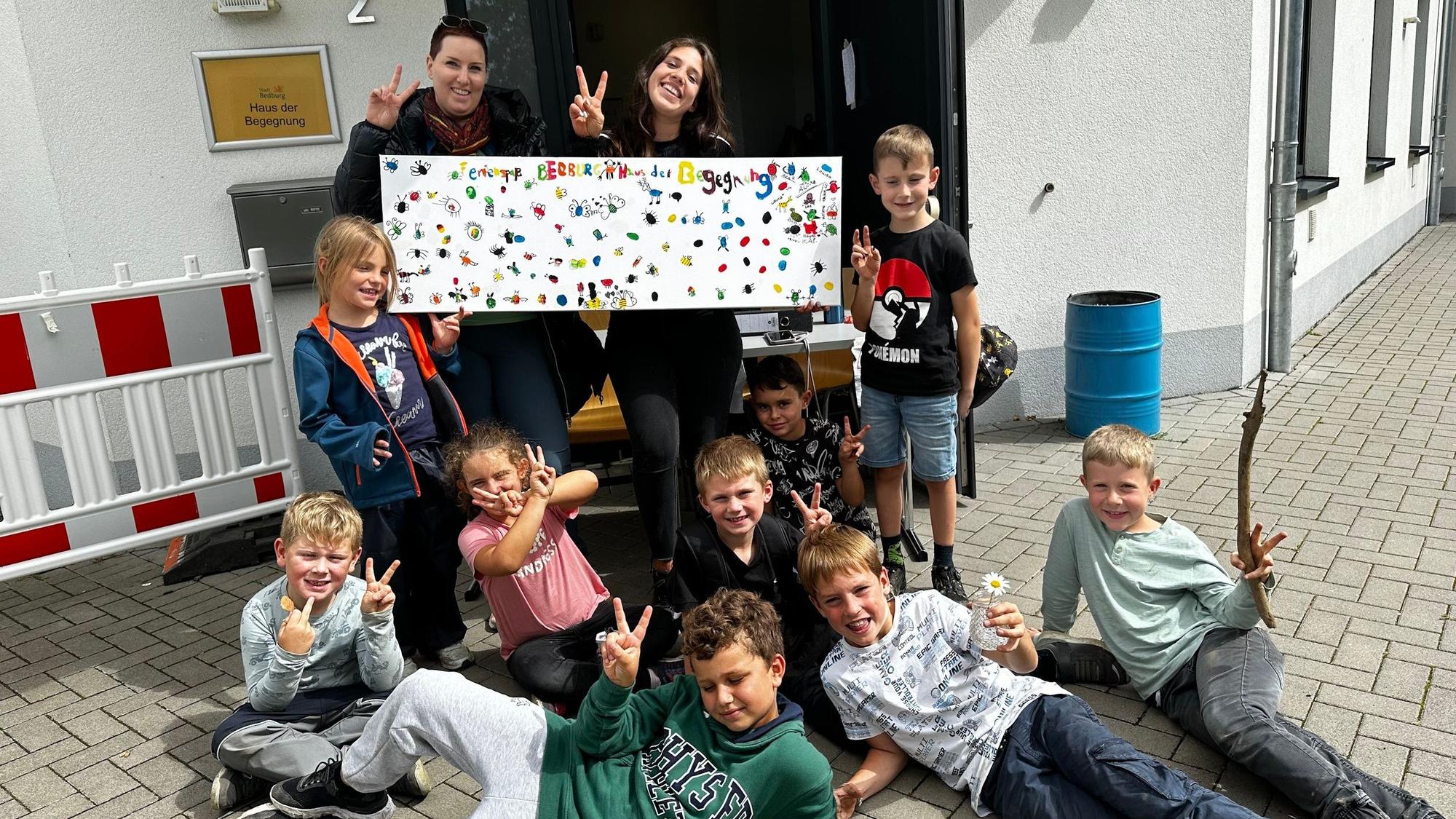 Zwei Frauen und mehrere Kinder posieren mit einem bunten Plakat vor einem Gebäude.