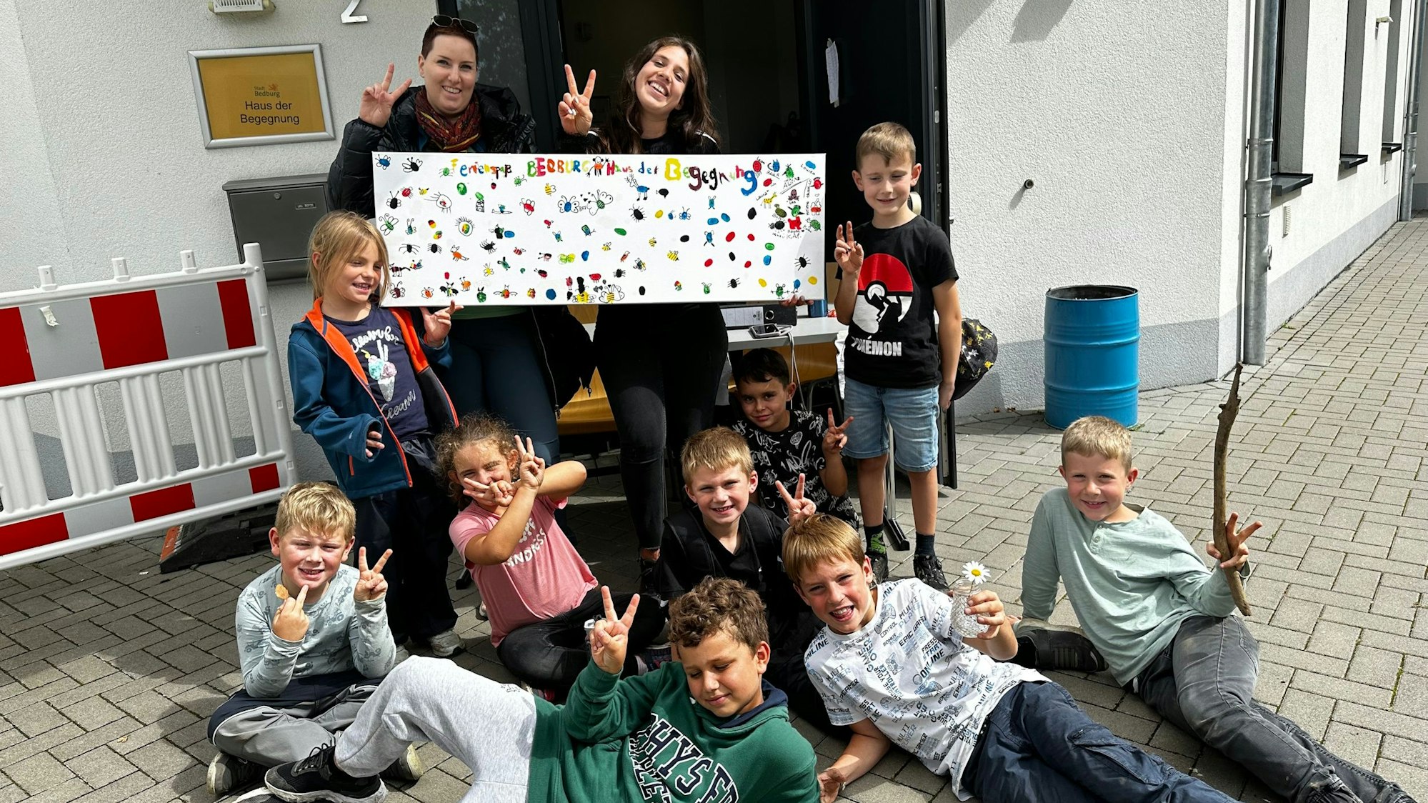 Zwei Frauen und mehrere Kinder posieren mit einem bunten Plakat vor einem Gebäude.