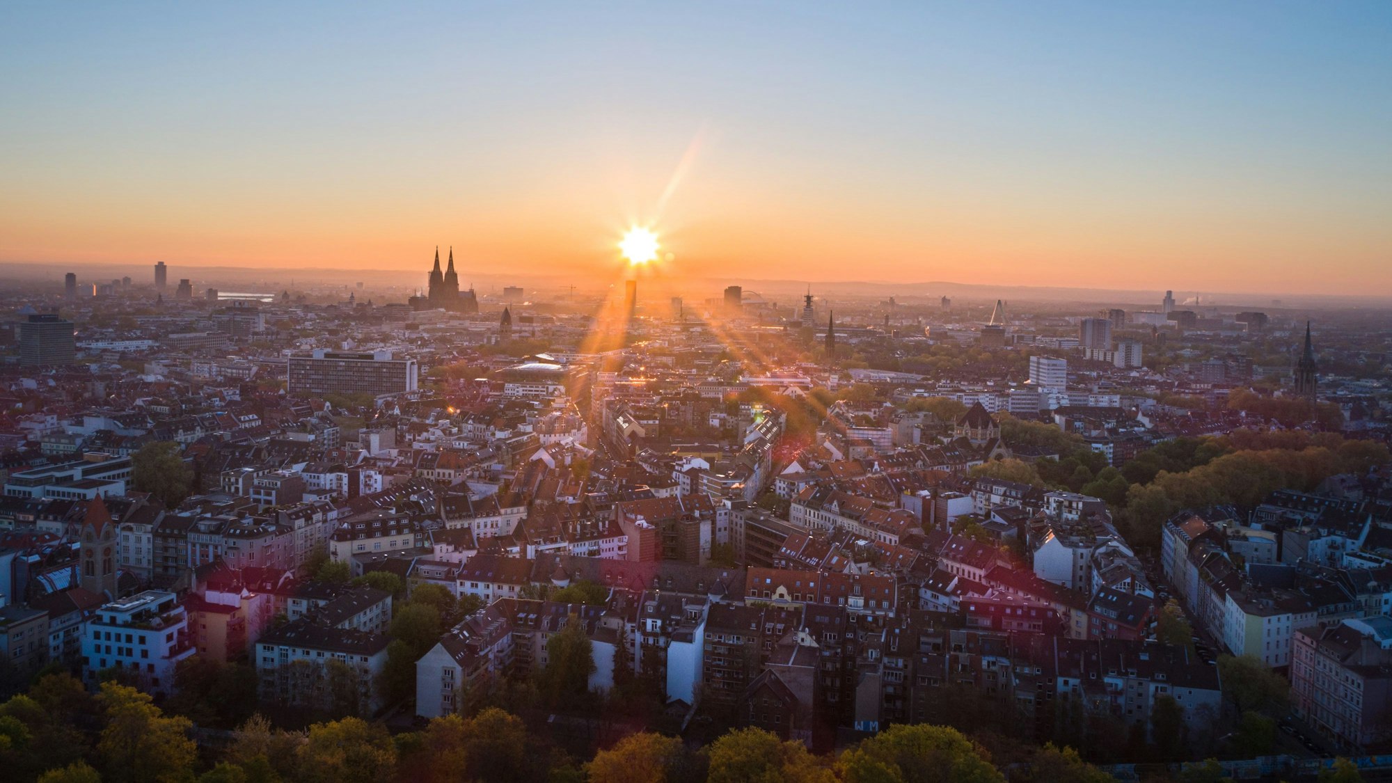 19.04.2017, Köln: Die Sonne geht an einem wolkenlosen Morgen mit blauem Himmel neben dem Kölner Dom (Hohe Domkirche St. Petrus) über dem Stadtzentrum auf. Foto: Matthias Heinekamp (Luftaufnahme mit Drohne) - Sonnenaufgang, Luftbild - Großstadt, Sunrise, Wetter, Innenstadt