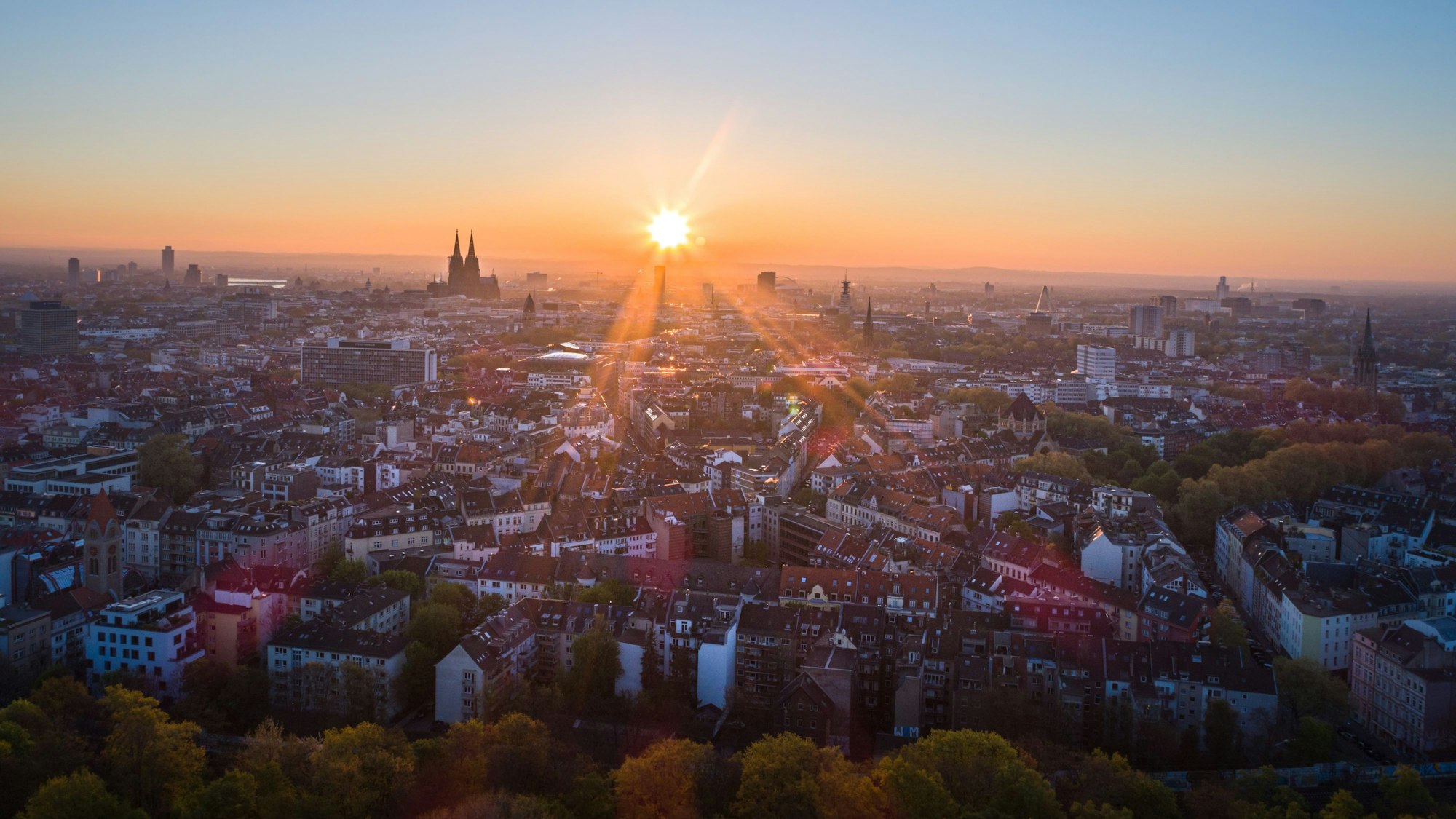 19.04.2017, Köln: Die Sonne geht an einem wolkenlosen Morgen mit blauem Himmel neben dem Kölner Dom (Hohe Domkirche St. Petrus) über dem Stadtzentrum auf. Foto: Matthias Heinekamp (Luftaufnahme mit Drohne) - Sonnenaufgang, Luftbild - Großstadt, Sunrise, Wetter, Innenstadt