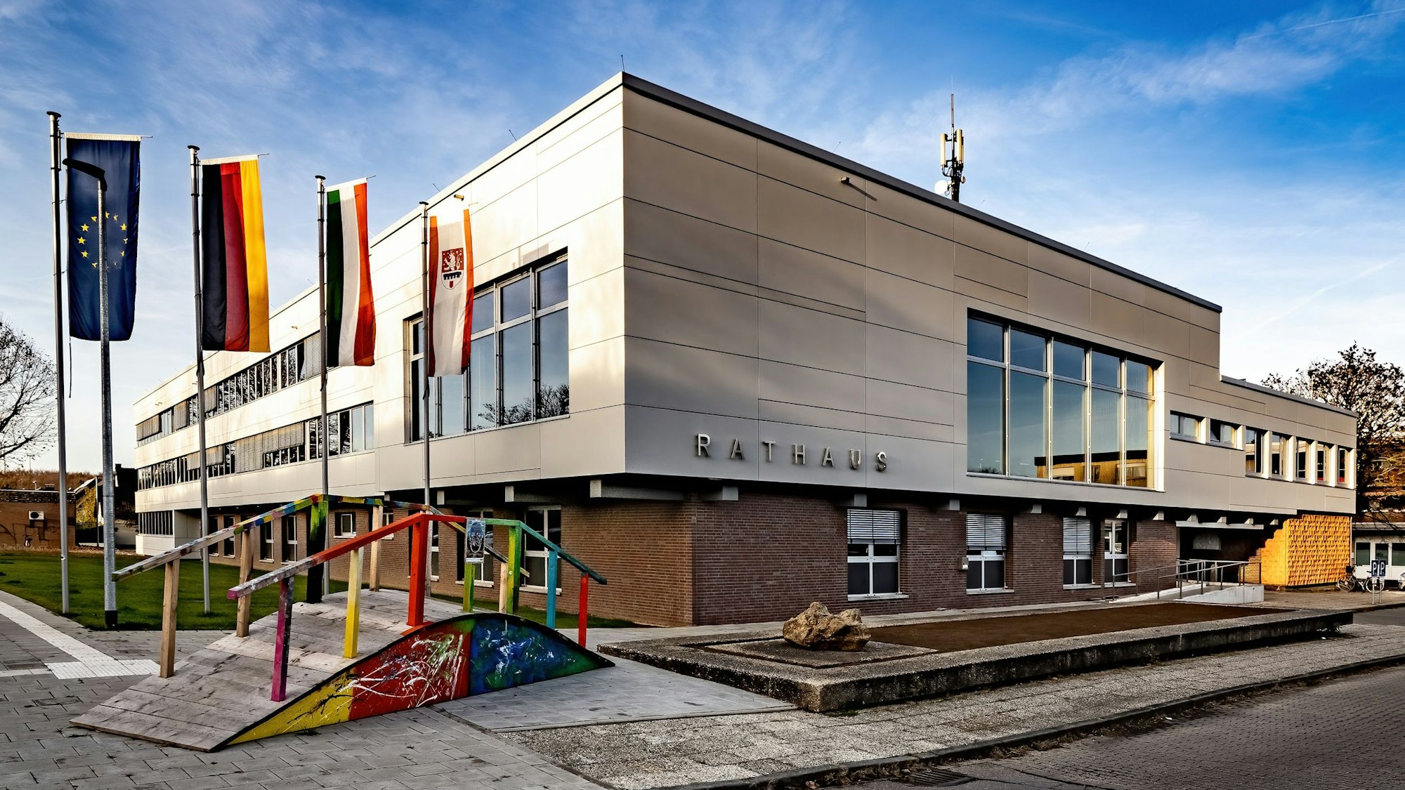 Vor einem großen würfelförmigen Bau wehen Flaggen an hohen Masten und steht eine bunt bemalte hölzerne Brücke.
