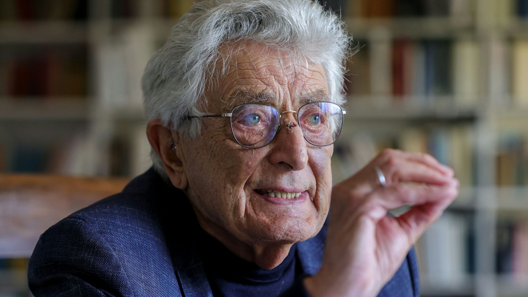 Gerhart Baum sitzt in seiner Wohnung vor einem Bücherregal und gestikuliert mit der rechten Hand.