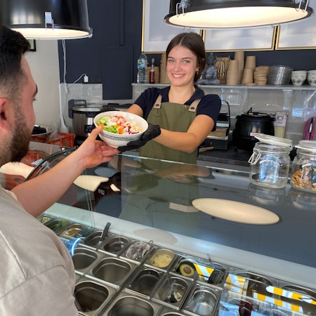 Das Foto zeigt eine Mitarbeiterin, die einem Kunden eine Schüssel mit Salat und Gemüse, eine sogenannte Bowl, anreicht.