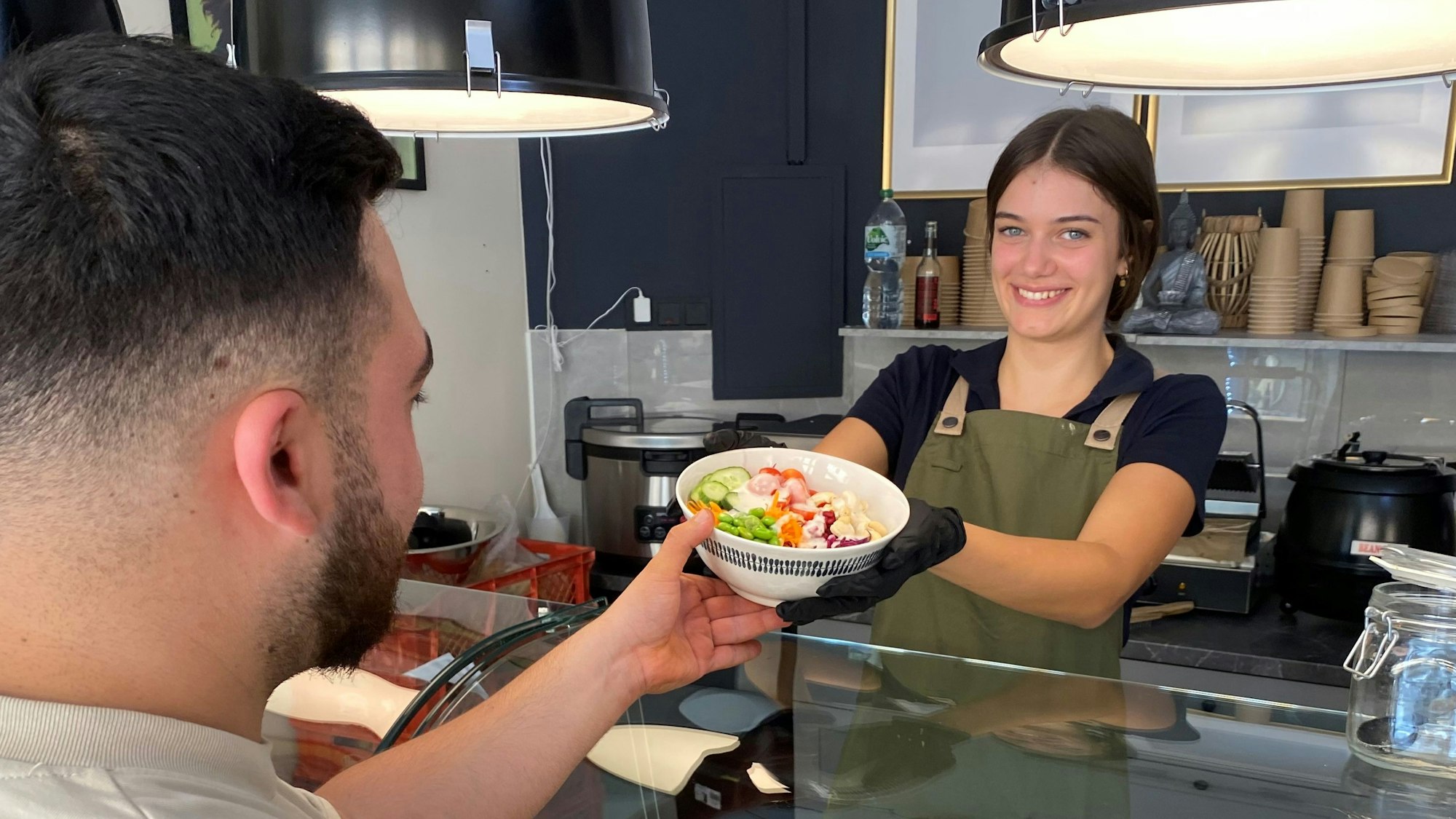 Das Foto zeigt eine Mitarbeiterin, die einem Kunden eine Schüssel mit Salat und Gemüse, eine sogenannte Bowl, anreicht.