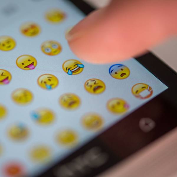 Eine Frau tippt auf das Display eines Smartphones, auf dem zahlreiche Emojis in der Nachrichten-App Whatsapp zu sehen sind.