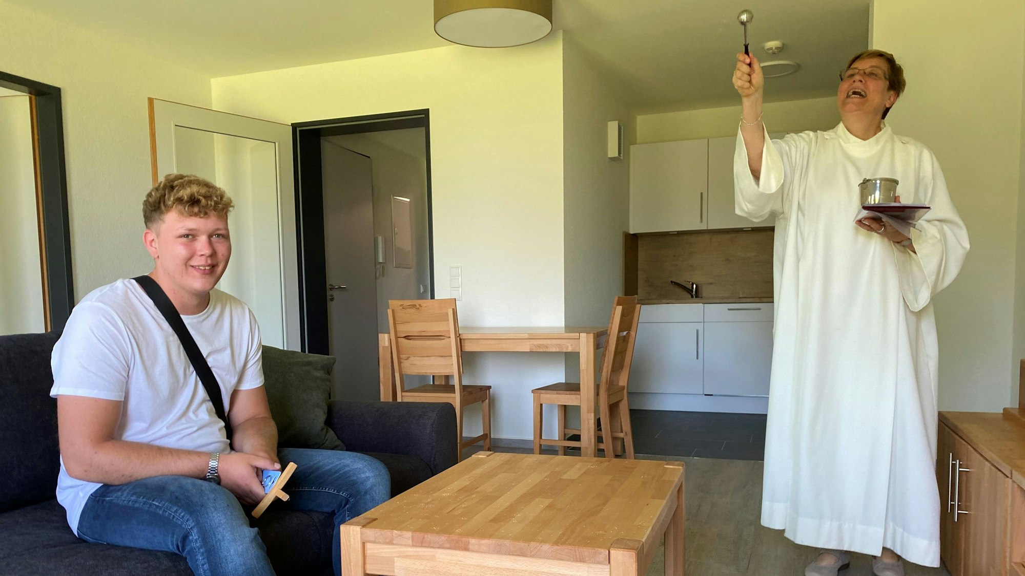 Seelsorgerin Dorothea Grimm steht in der noch kahlen Wohnung von Cevin Wego. Mit einem Weihwasserwedel segnet sie das Zimmer. Cevin Wego sitzt auf der Couch.
