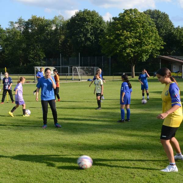Auf einem Trainingsplatz machen Mädchen Fußball-Übungen. Sie tragen blaue, gelbe und weiße Trikots.