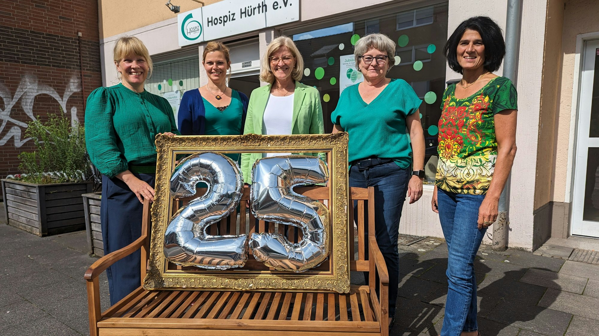Vier Frauen stehen hinter einer Bank, auf der ein Rahmen mit der silbernen Zahl 25 platziert ist.