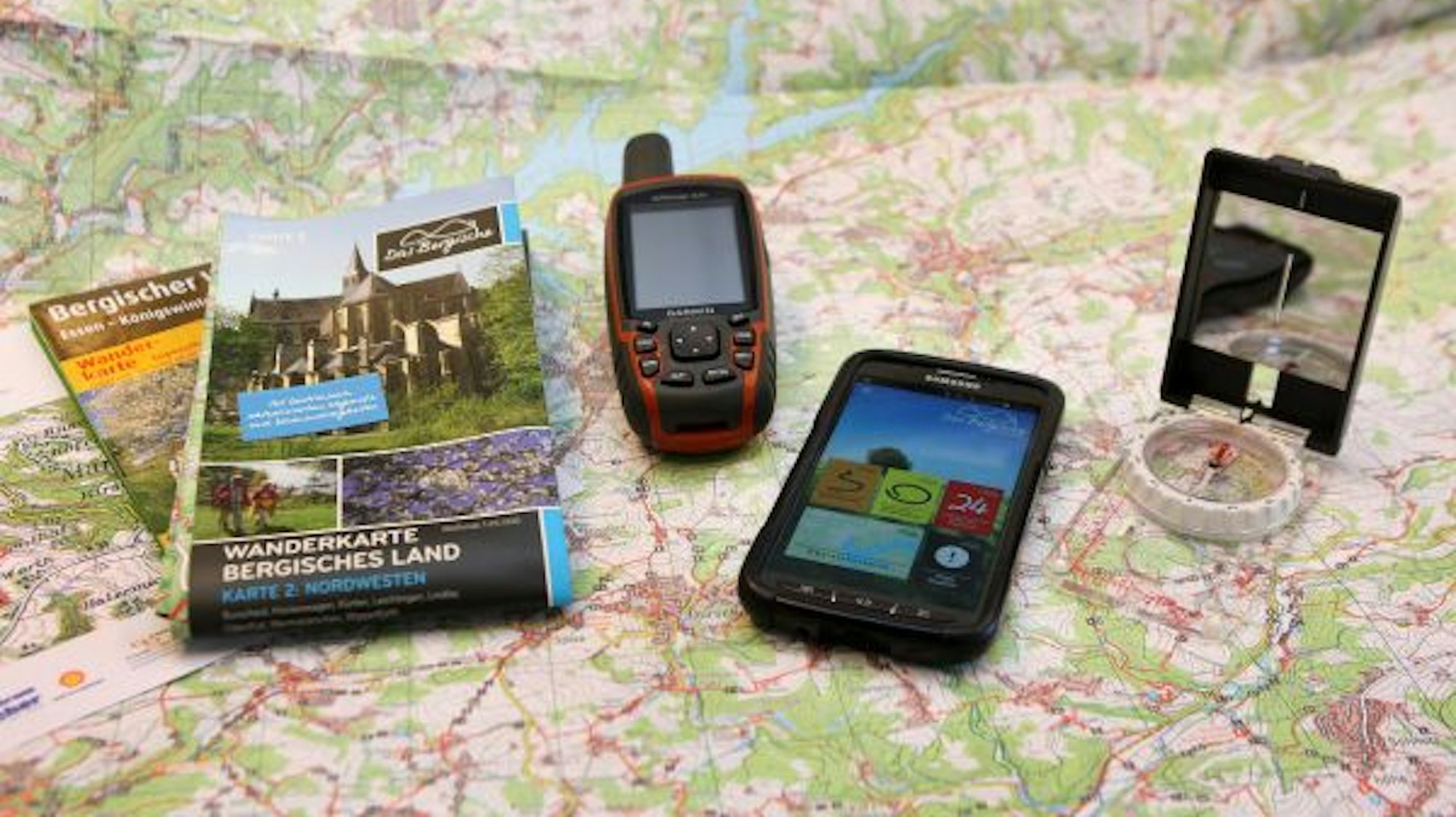 Eine Wanderkarte, ein Navigationsgerät, ein Handy mit Wander-App und ein Kompass liegen auf einer Wanderkarte.
