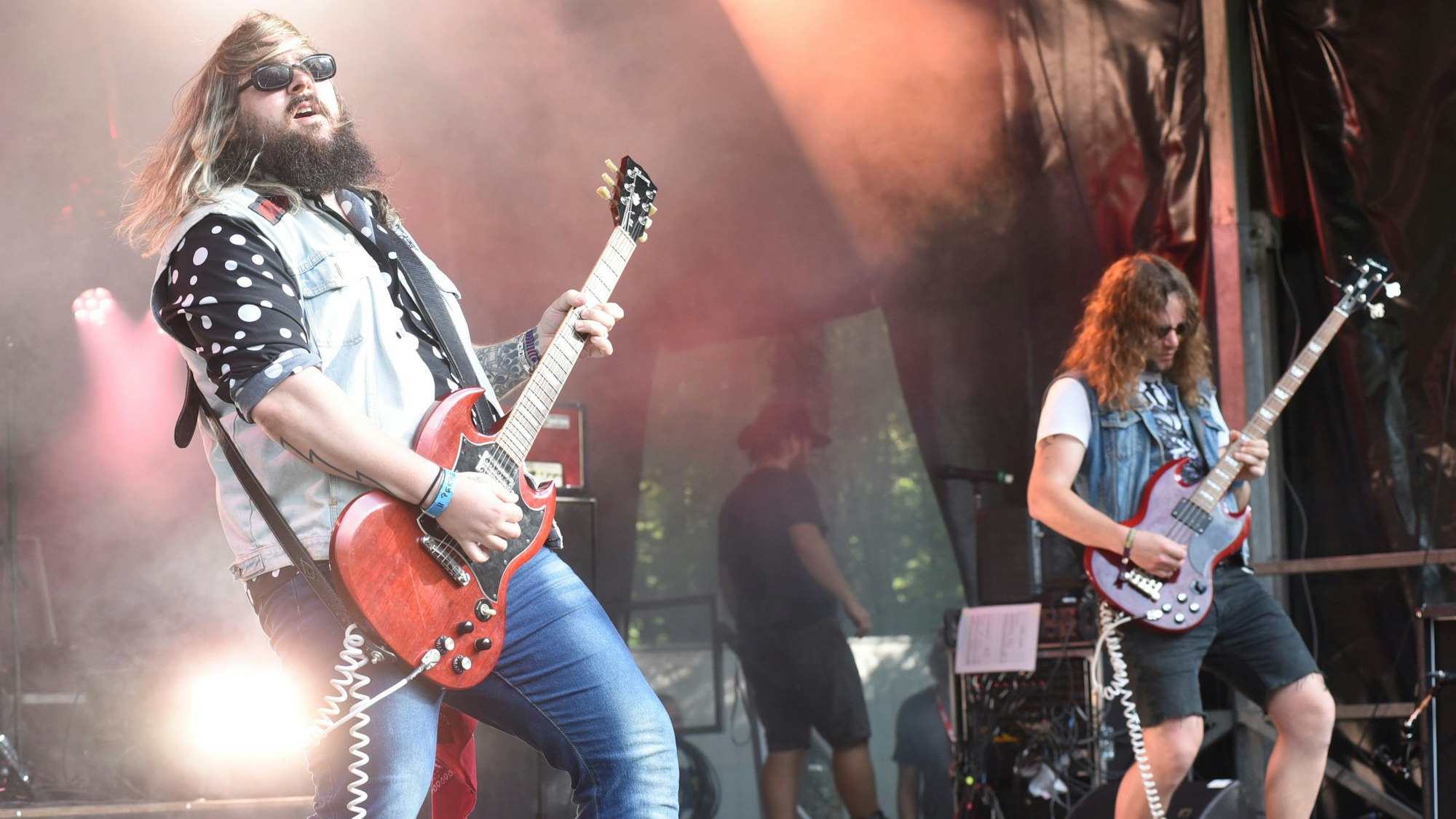 Zwei Musiker mit wehenden, langen Haaren spielen auf ihren Instrumenten auf einer von Scheinwerfern beleuchteten Bühne.