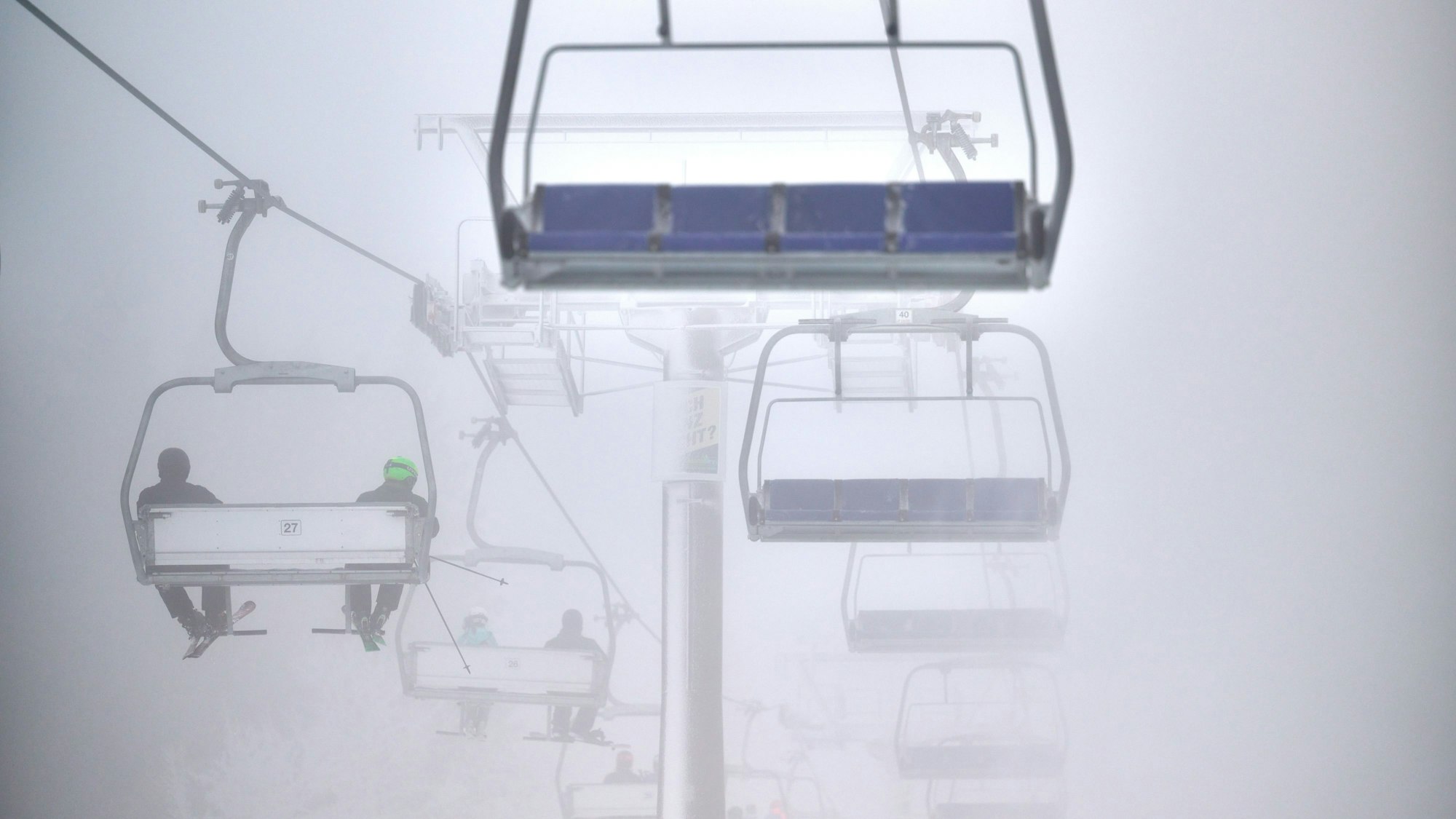 Wintersportler fahren mit einem Sessellift über eine künstlich beschneite Piste. Es ist neblig. (Symbolbild)