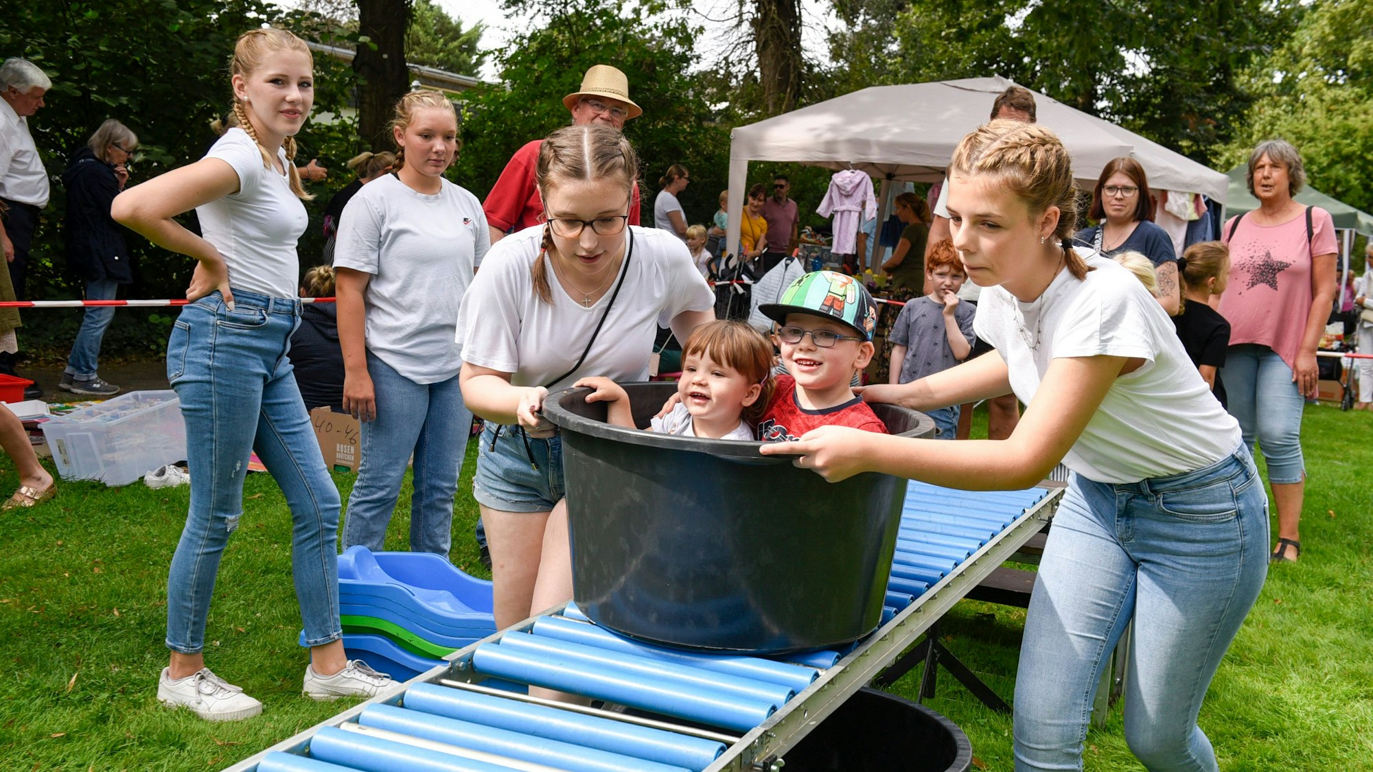 Das Bild zeigt kleine Kinder, die in einem Bottich sitzend von jugendlichen Mädchen, einheitlich bekleidet in weißem T-Shirt und Jeans, über eine Trockenrutschbahn geschoben werden.