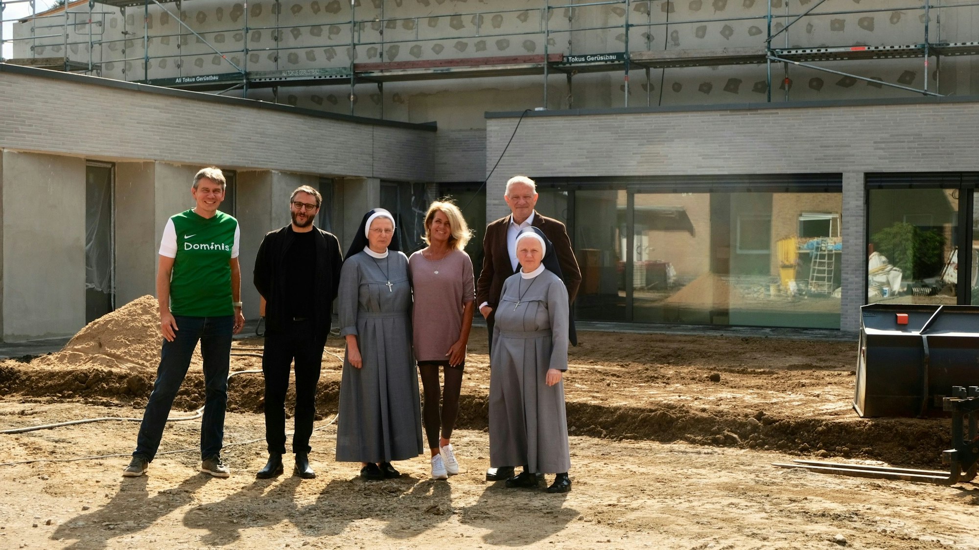 Drei Männer und drei Frauen, davon zwei im Habit der Ordensschwestern, stehen vor einem grau verklinkerten Neubau.