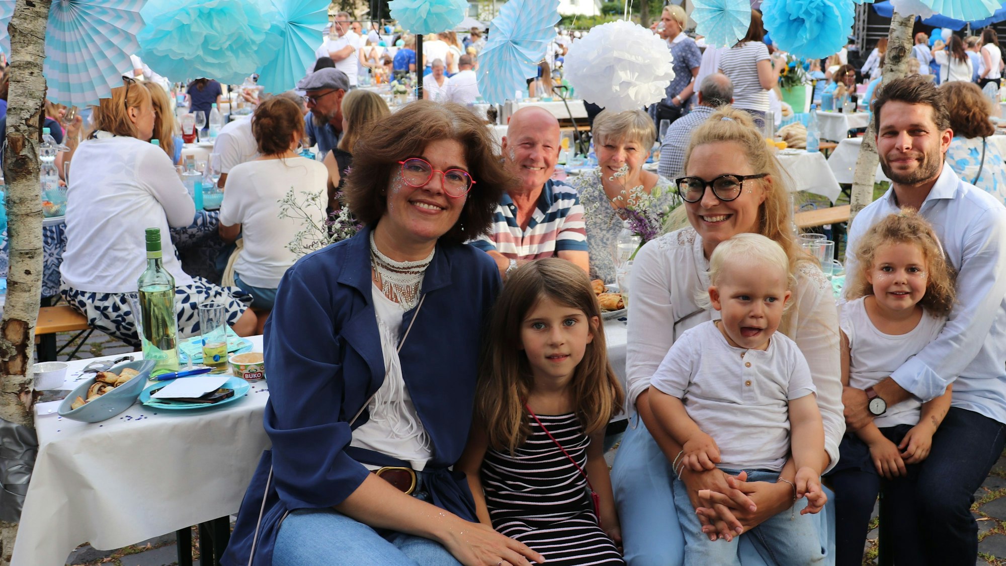 Zwei Frauen, ein Mann und drei Kinder sitzen an einem dekorierten Tisch. Über ihnen schweben türkis-farbene Fächer.