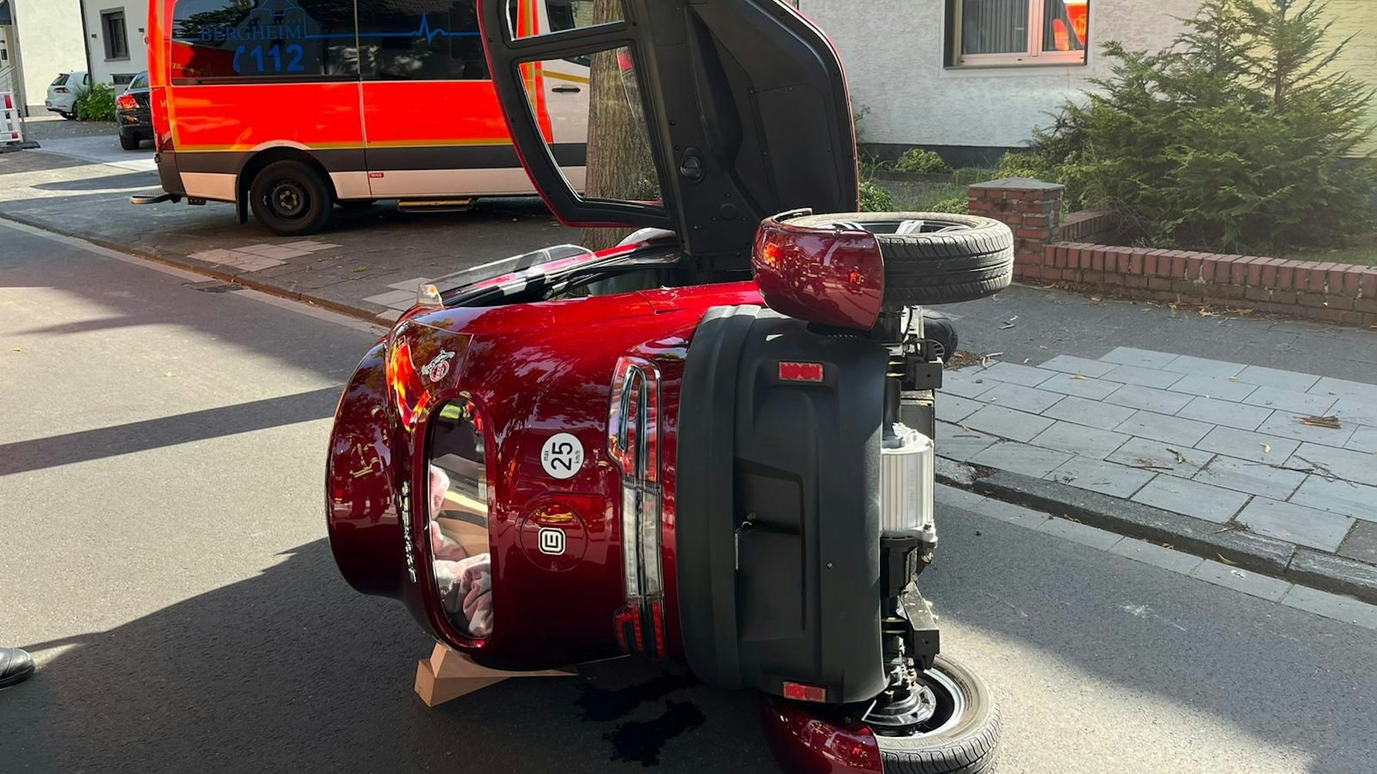 Der weinrote Kabinenroller liegt umgekippt auf der Straße, im Hintergrund steht ein Krankenwagen.