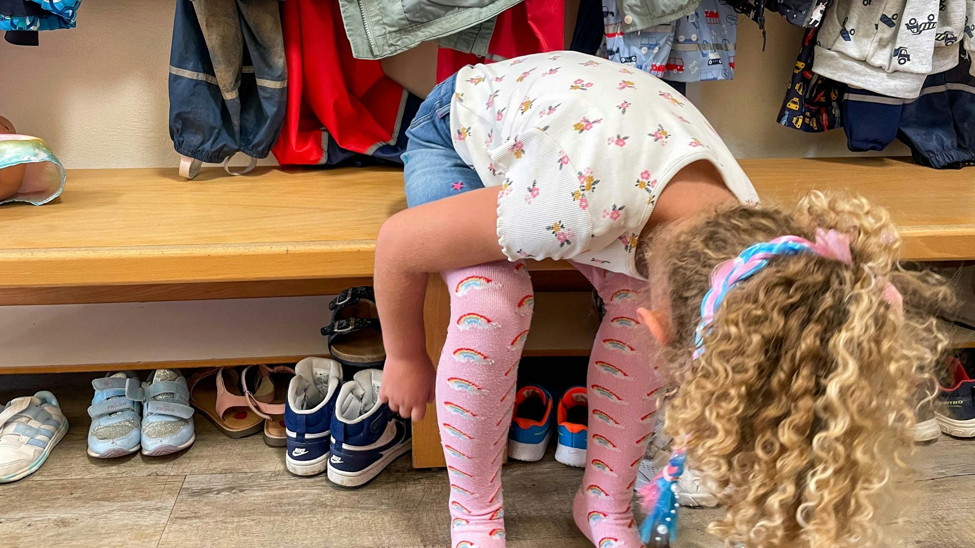 Das Bild zeigt ein Kindergartenkind, das sich auf einer Bank in der Kita die Schuhe anziehen will.