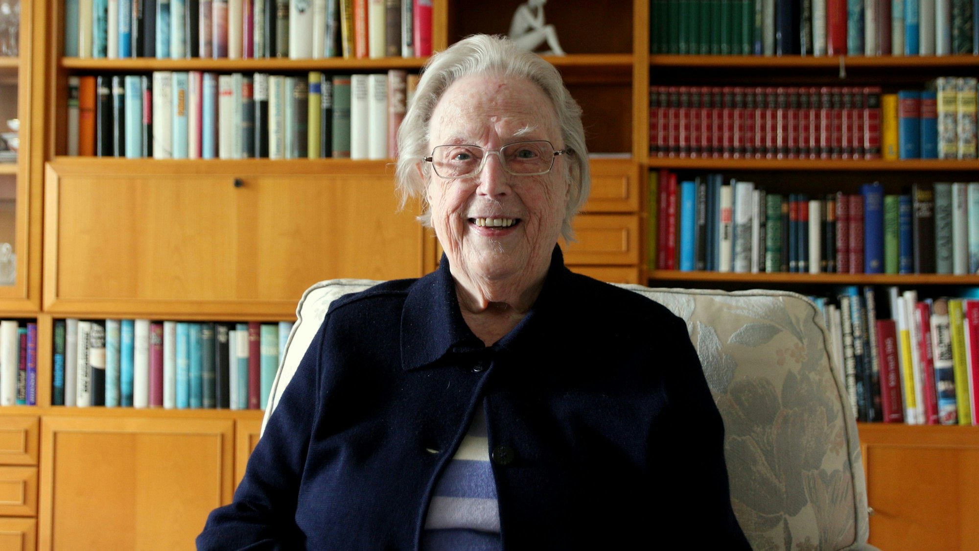 Eine lächelnde Frau sitzt in einem Sessel vor einer Bücherwand.