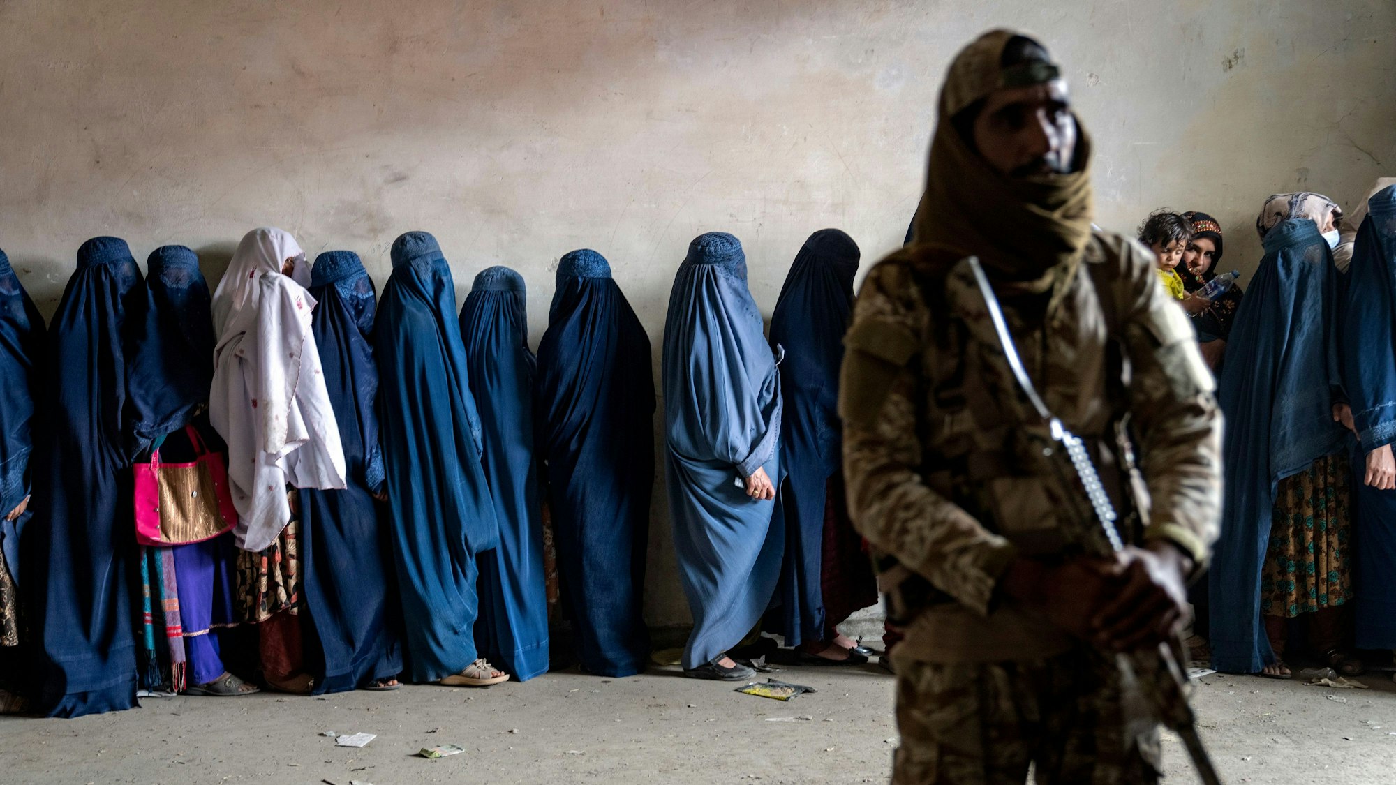 Afghanistan, Kabul: Ein Taliban-Kämpfer steht Wache, während Frauen darauf warten, von einer humanitären Hilfsorganisation verteilte Lebensmittelrationen zu erhalten.