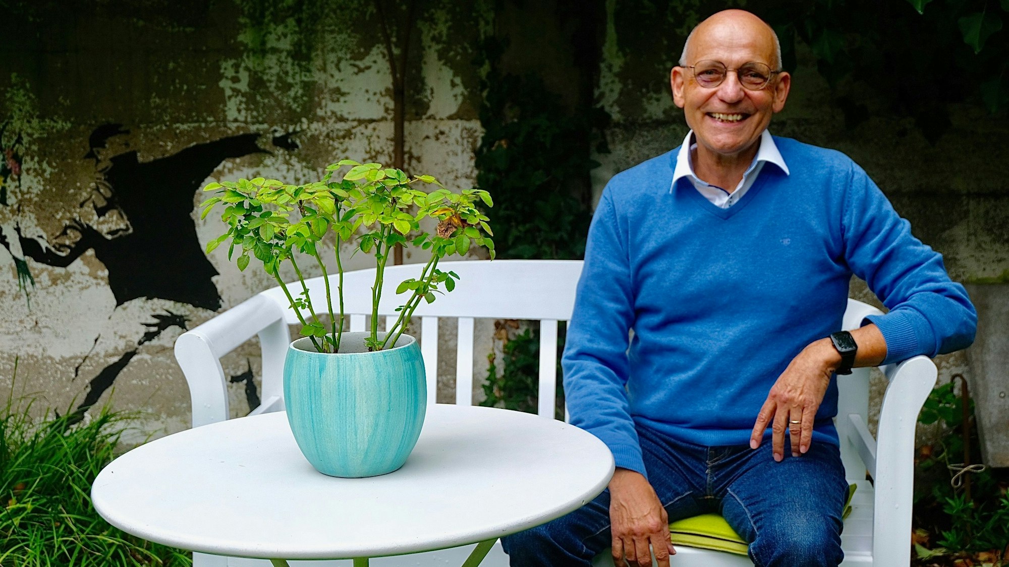 Ein braun gebrannter lachender Mann mit Glatze, Jeans und blauem Pulli sitzt auf einer weißen Bank an einem weißen Tisch, auf dem eine üppige Grünpflanze steht.