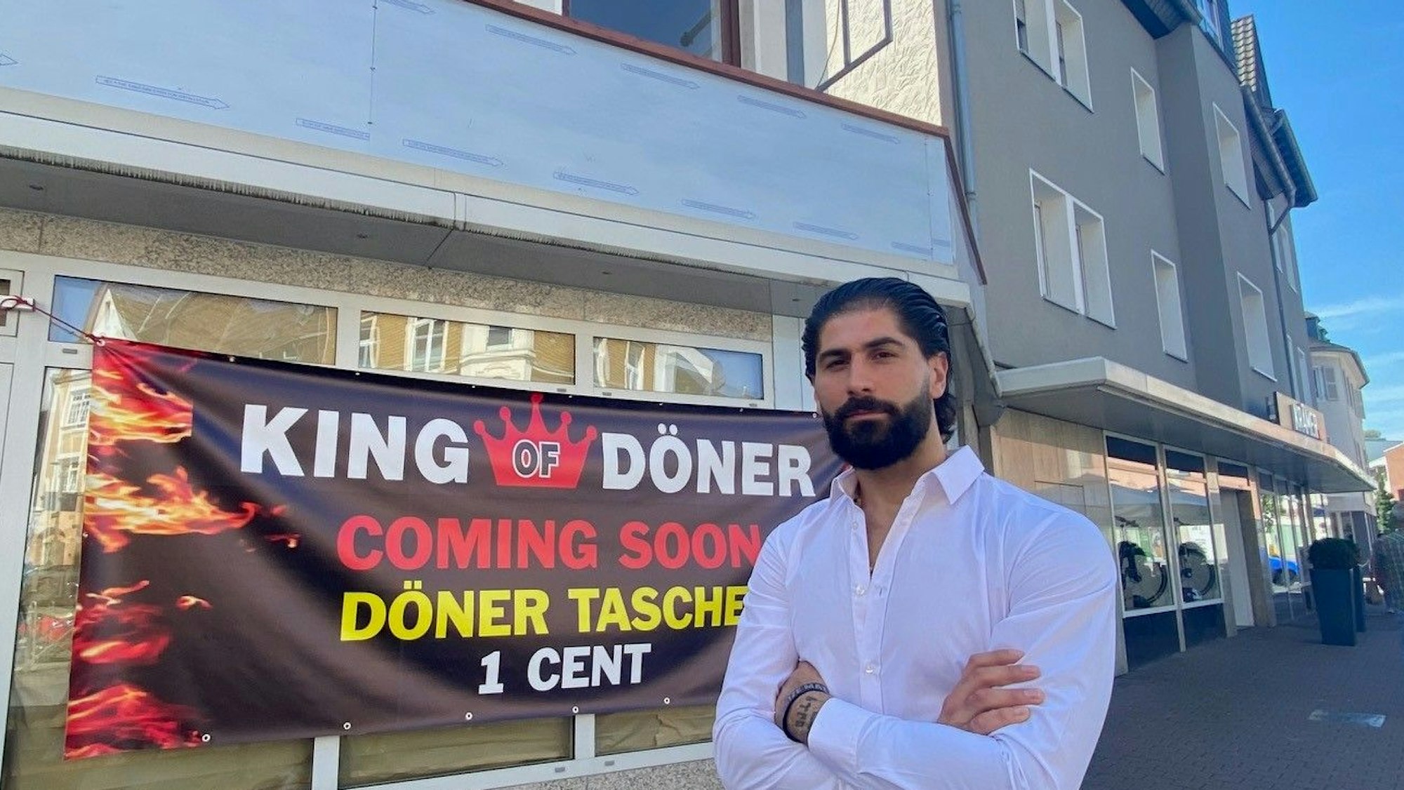 Das Bild zeigt Murat Gümüs bekleidet mit einem weißen Hemd vor seinem künftigen Restaurant. Ein Banner am Schaufenster kündigt zur Eröffnung Döner für einen Cent an.