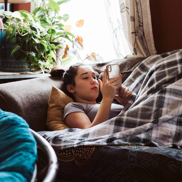 Eine Jugendliche, die unter einer Decke auf der Couch liegt und etwas auf dem Handy sieht.