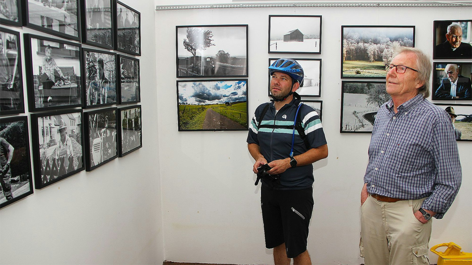 Dietrich Schubert in Straßen- und Erik Pütz in Radlerkleidung betrachten Fotografien an der Wand.