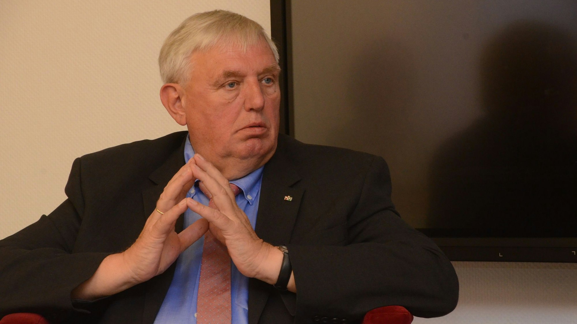 NRW-Minister Karl-Josef Laumann sitzt in einem kleinen roten Sessel. Er trägt ein blaues Hemd und eine altrosa Krawatte. Er hat die Fingerspitzen seiner Hände aufeinandergelegt.