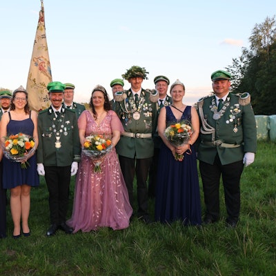 Mehrere Schützen in Uniform und Frauen in Ballkleidern stehen als Gruppe auf einer Wiese zusammen. Im Hintergrund ist die Fahre der Schützen aus Lieberhausen zu sehen.
