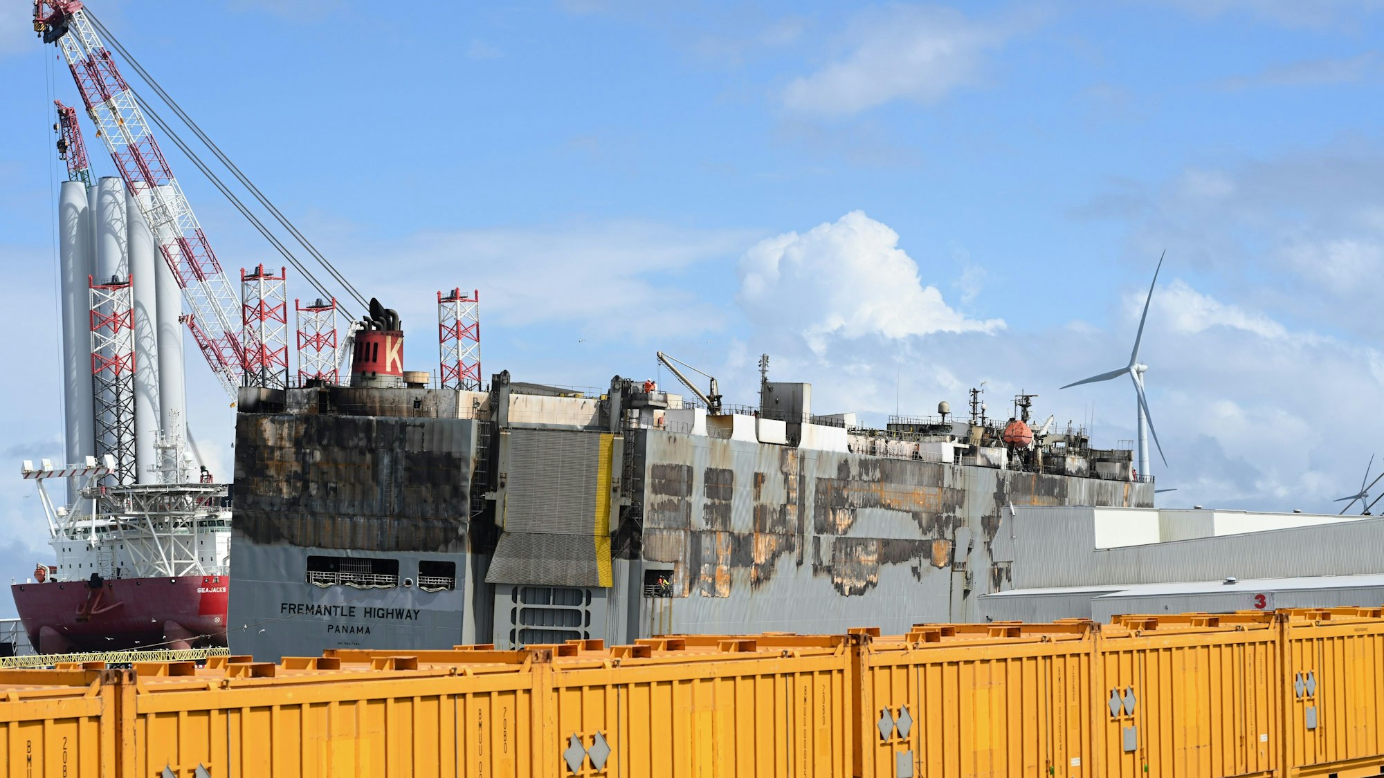 Der schwer beschädigte Frachter „Fremantle Highway“ liegt im Hafen von Eemshaven an der niederländischen Nordseeküste. Sie wird von einem Notfallschlepper gestützt. Davor sind gelbe Container aufgebaut.