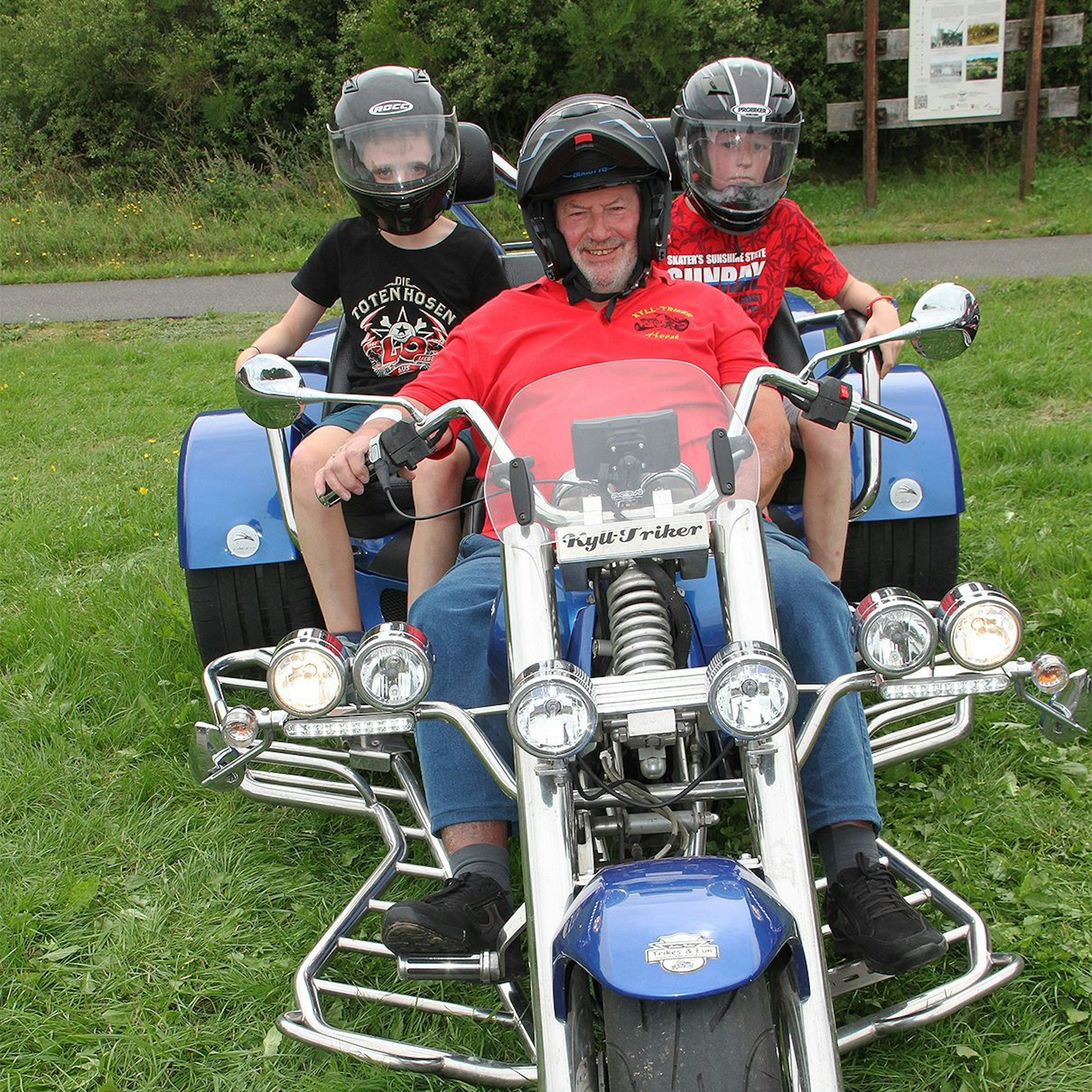 Auf einem Trike – einem Motorrad mit drei Rädern – sitzt ein Erwachsener mit zwei Kindern.