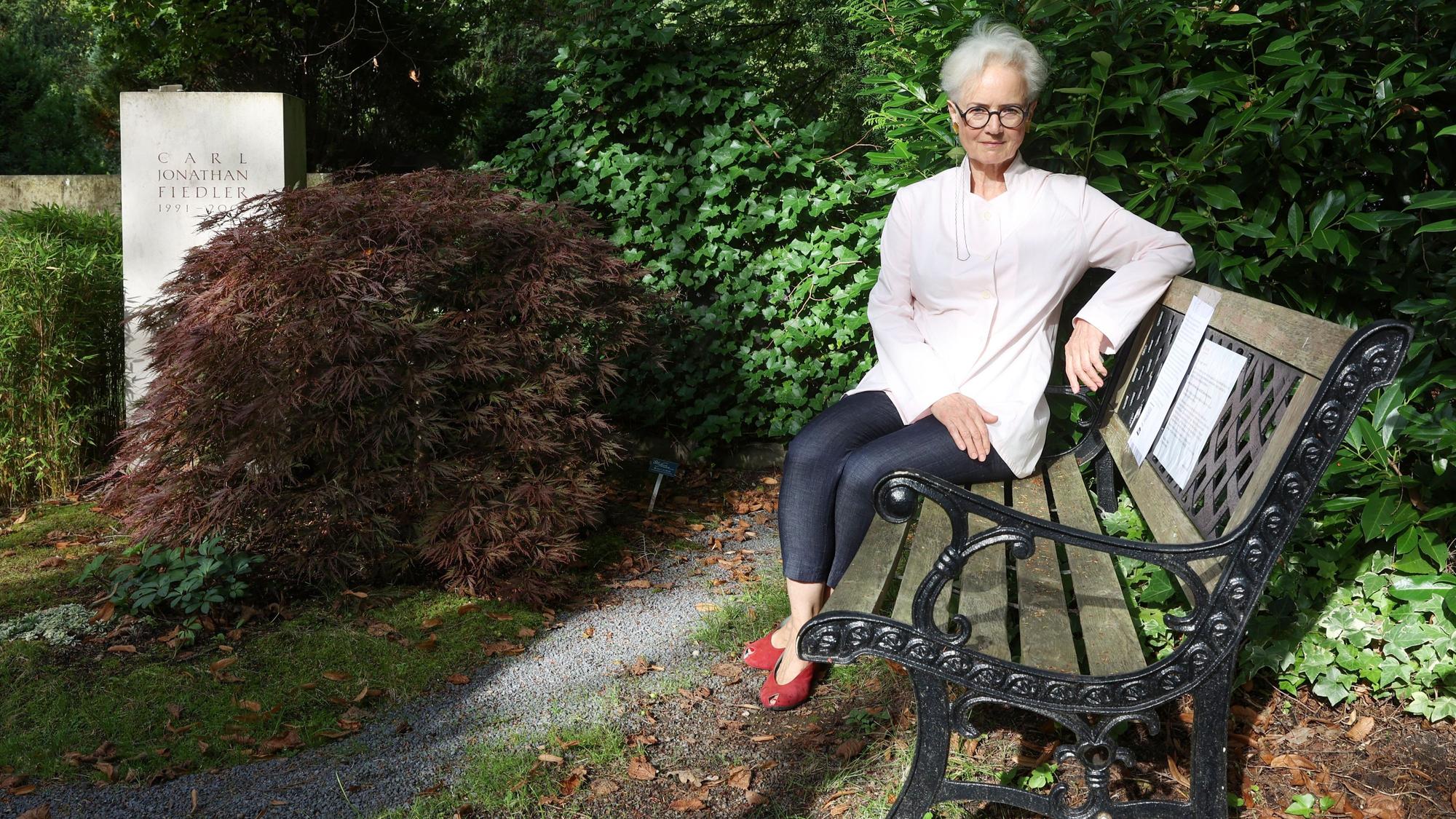 Dorothee Fiedler sitzt auf einer Bank aus Holz mit einem Gestell aus Metall. Neben ihr ist das Grab ihres Sohnes.