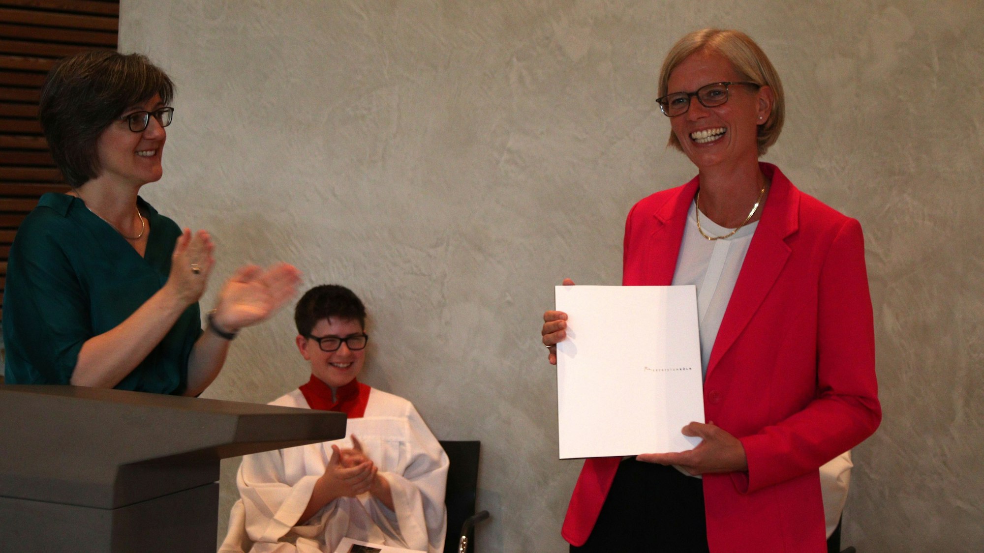 Schulleiterin Carolin Neswadba hält ihre Urkunde, Bernadette Schwarz-Boenneke applaudiert. Ein Messdiener im Hintergrund freut sich.