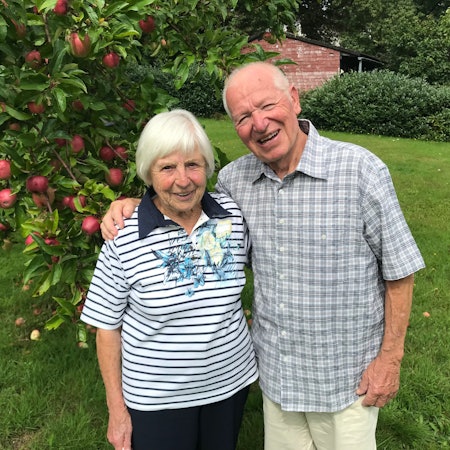 Ein älteres Ehepaar steht Arm in Arm vor einem Apfelbaum.