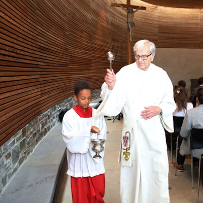 Diakon Dr. Ulrich Günzel segnet die Schulkapelle des St.-Angela-Gymnasiums ein. Neben ihm hält ein Messdiener ein Weihwasserfässchen.