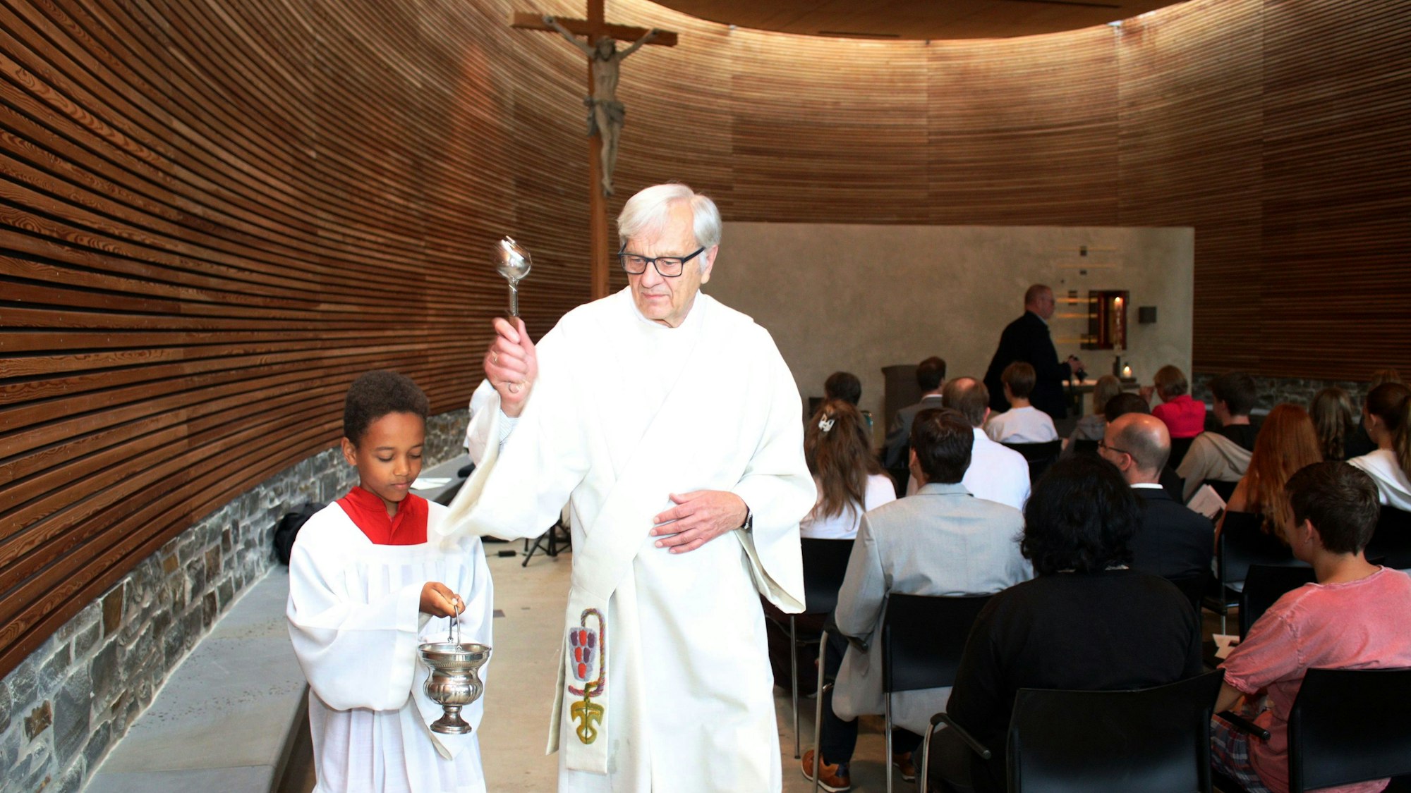 Diakon Dr. Ulrich Günzel segnet die Schulkapelle des St.-Angela-Gymnasiums ein. Neben ihm hält ein Messdiener ein Weihwasserfässchen.
