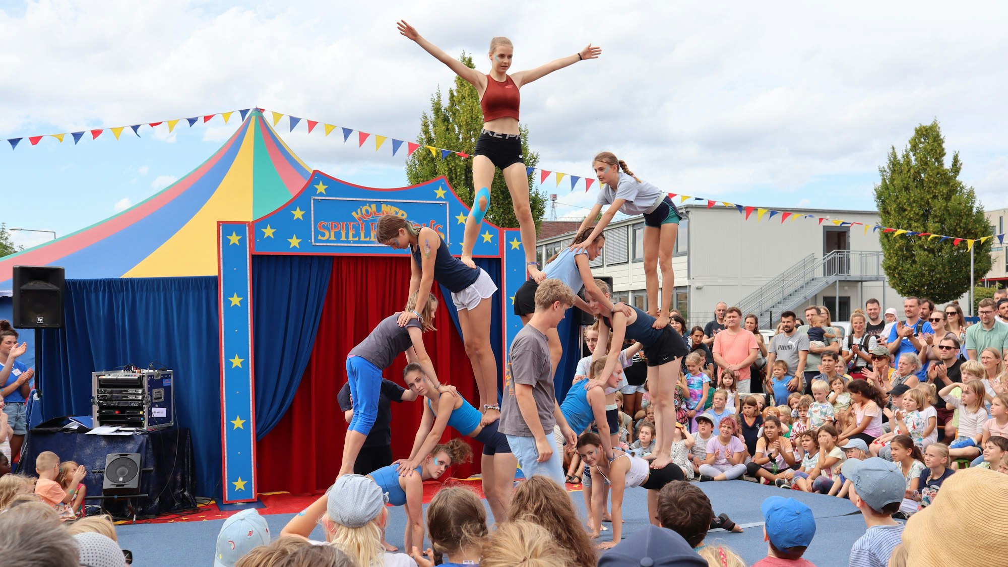 Bunte Zelte stehen auf Parkplatzflächen, es gibt Bäume und ein rotes Bürogebäude.